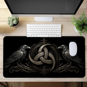 Op de afbeelding: Een rechthoekige zwarte bureaumat met een centraal triquetra-symbool in metaaltinten, omringd door ingewikkeld vlechtwerk. Twee gedetailleerde zwarte raven bevinden zich aan weerszijden van het embleem, zittend op donkere, gevlochten takken.