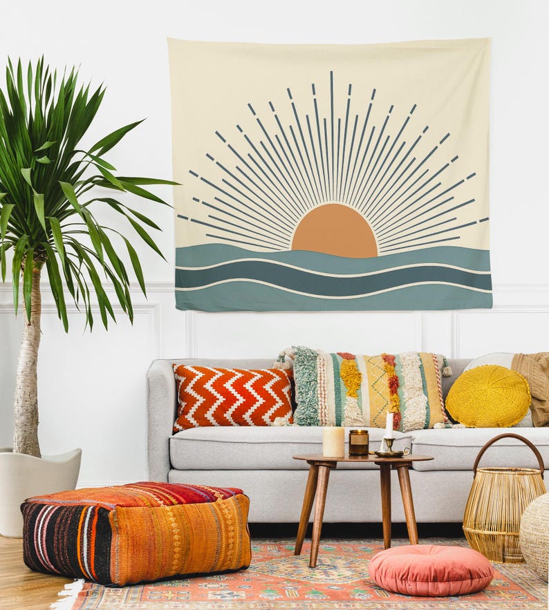 Pu&ograve; includere: Un arazzo da parete beige con un design a sole e oceano. Il sole &egrave; arancione, con raggi blu scuro. L'oceano &egrave; rappresentato da onde blu. L'arazzo &egrave; appeso sopra un divano grigio con cuscini colorati e un pouf fantasia.