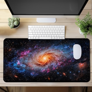 Könnte beinhalten: Eine große, rechteckige Schreibtischunterlage mit einem lebendigen Galaxy-Design in wirbelnden Orange-, Blau- und Lilatönen. Eine weiße Maus und Tastatur liegen auf dem Holzschreibtisch. Ein Computermonitor und Topfpflanzen sind ebenfalls zu sehen.