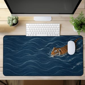 May include: A rectangular desk mat featuring a detailed illustration of an orange and black tiger swimming through deep blue water with white stylised wave patterns. The mat is positioned on a light wood surface beneath a computer monitor and keyboard.