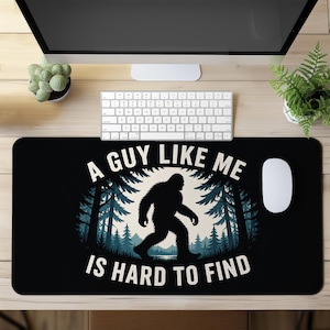 May include: Black desk mat featuring a Bigfoot silhouette walking through a forest scene. The text "A GUY LIKE ME IS HARD TO FIND" is printed in white. The mat is rectangular and sits on a wooden desk.