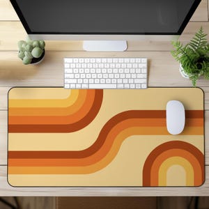 Puede incluir: Una alfombrilla de escritorio rectangular con un patrón repetitivo de rayas curvas y suaves en tonos amarillo, naranja y marrón oscuro sobre un fondo crema claro. La alfombrilla está colocada sobre una superficie de madera bajo un teclado y ratón.