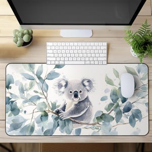 May include: A rectangular mouse mat with a watercolour illustration of a koala bear on a branch with blue-green eucalyptus leaves. The mat has a white background and a black border. A white mouse and keyboard are on the wooden desk.