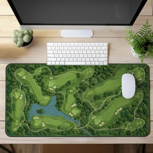 May include: A rectangular desk mat featuring a detailed top-down aerial illustration of a sprawling golf course with lush green fairways, sand bunkers, dense patches of trees, and a winding blue water hazard spanning across the center.