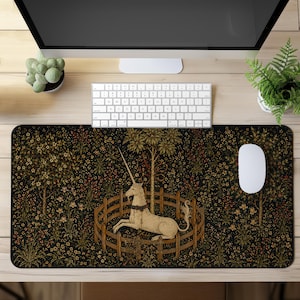 May include: A rectangular desk mat featuring a detailed reproduction of The Unicorn in Captivity tapestry. The design displays a cream-coloured unicorn resting inside a wooden fence, surrounded by a dark background filled with colourful flowers.