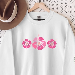 May include: White sweatshirt with a pink hibiscus flower design. The sweatshirt is hanging on a hanger against a white background with a black geometric pattern.