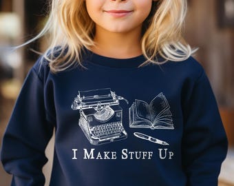 May include: Navy blue sweatshirt featuring a white illustration of a typewriter, open book, and pen. The text "I MAKE STUFF UP" is printed below the graphic. The sweatshirt is worn by a young person with blonde hair.