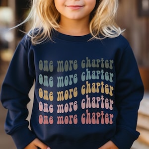 May include: A navy blue sweatshirt with the text "one more chapter" repeated five times in a rainbow gradient. The text is in a retro font style.