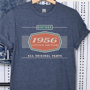 May include: Heather navy t-shirt with the text "VINTAGE 1956 LIMITED EDITION ALL ORIGINAL PARTS" in white and orange. The shirt is hanging on a wooden hanger. The background includes a pair of blue jeans and a black and white patterned wall.
