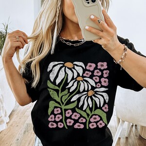K&ouml;nnte beinhalten: Schwarzes T-Shirt mit einem grafischen Druck von wei&szlig;en G&auml;nsebl&uuml;mchen und rosa Blumen. Das Shirt ist oversized und hat eine lockere Passform.