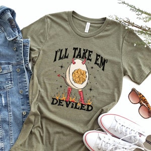 May include: Heather olive green t-shirt with the text "I'LL TAKE EM' DEVILED" and a cartoon deviled egg character with horns, a tail, and boots. A denim jacket, white sneakers, and leopard print sunglasses are also in the image.