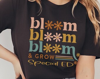 Puede incluir: Una camiseta negra con un diseño gráfico que dice "bl**m bl**m bl**m & GROW Special ED*" en letras de colores con acentos de flores.