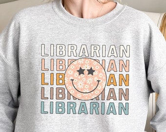 Peut inclure: Un sweat-shirt gris avec le mot "LIBRARIAN" répété cinq fois dans un design empilé. Un visage souriant à motif de marguerite avec des yeux en forme d'étoile est centré entre les mots.