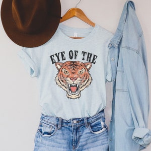 May include: A light blue t-shirt with a tiger graphic and the text "Eye of the Tiger". The shirt is on a hanger with a brown hat and a light blue denim jacket.