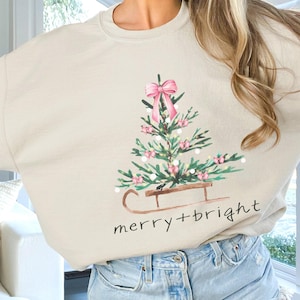 Könnte beinhalten: Ein beiger Pullover mit einem Weihnachtsbaum-Motiv und dem Text "merry+bright".