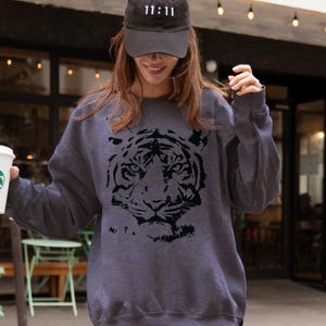 Op de afbeelding: Donkergrijs sweatshirt met een zwart tijgergezicht ontwerp. De persoon draagt ​​zwarte leggings en een zwarte baseball cap met de tekst "11:11". De foto is genomen op een zebrapad.