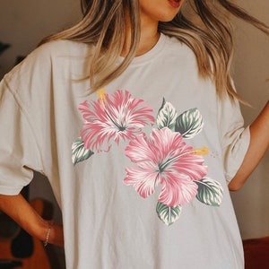 Puede incluir: Una camiseta blanca de gran tama&ntilde;o con estampado floral rosa y verde. Las flores son hibiscos.