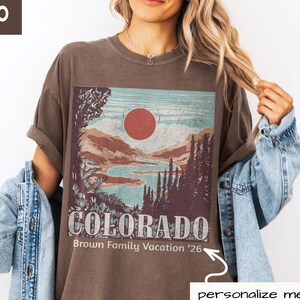 May include: Espresso-colored t-shirt featuring a Colorado landscape design with a red sun, mountains, and a lake. The text "COLORADO" and "Brown Family Vacation '26" are printed on the front. The model is wearing a denim jacket.