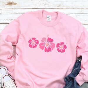 May include: A pink sweatshirt with a row of three pink hibiscus flowers printed on the front. The sweatshirt is laying on a white wooden surface with a pair of white sneakers and blue jeans.