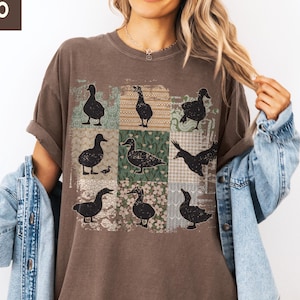 May include: Espresso-colored t-shirt featuring a patchwork design with nine black duck silhouettes. The ducks are set against a backdrop of floral and patterned squares. The shirt has short sleeves and a relaxed fit.
