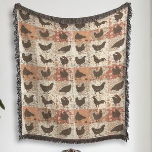 May include: A woven throw blanket featuring a pattern of brown chickens on a beige and orange background. The blanket is hanging on a white wall above a white sofa.