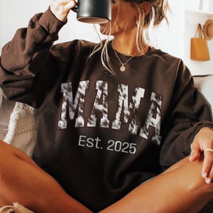 May include: Dark chocolate-colored sweatshirt with the word "MAMA" in a cow print design and the text "Est. 2025". A black mug is held up to the face. The sweatshirt is worn with fuzzy slippers.