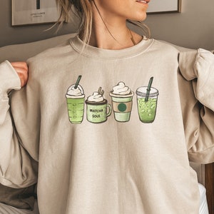 May include: A beige sweatshirt with a graphic of four green iced coffee drinks. The text "Matcha Soul" is printed on the sweatshirt.
