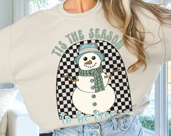 May include: A light beige sweatshirt with a graphic of a snowman in a checkered arch. The text "Tis the season to be freezin'" is printed on the sweatshirt.