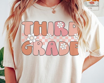 Op de afbeelding: Een wit T-shirt met de tekst "THIRD GRADE" in roze en oranje letters met witte madeliefjes en een geruit patroon. Het shirt wordt gedragen door een model. De tekst "IVORY" staat in de rechterbenedenhoek van de afbeelding.
