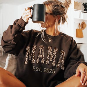 Puede incluir: Una sudadera de color chocolate oscuro con la palabra "MAMA" en letras con estampado de leopardo y "Est. 2025" impreso debajo. Una taza negra se sostiene frente a la cara. La sudadera es parte de una colección de ropa de estar por casa.