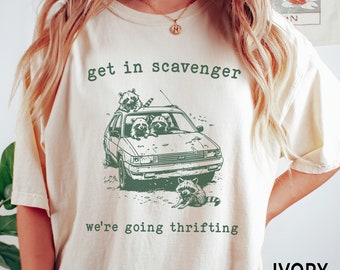 Może przedstawiać: Kremowa koszulka z zieloną grafiką szopów w samochodzie, z napisem "get in scavenger we're going thrifting". Koszulka ma luźny krój.