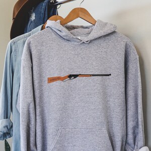 May include: A gray hooded sweatshirt with a black and brown rifle graphic printed on the front. The hoodie has a kangaroo pocket and a drawstring hood.