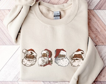 May include: A beige sweatshirt with a row of four black Santa Claus faces wearing red hats. The Santa Claus faces are in a row across the center of the sweatshirt.