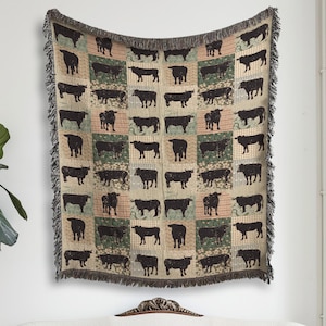 May include: A woven throw blanket featuring a repeating grid pattern of black cow silhouettes against alternating squares of beige, green, and orange backgrounds. The blanket has a fringed edge and is displayed hanging on a white wall.