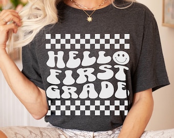 May include: A dark grey heather t-shirt with a white graphic that says "Hello First Grade" in a retro font with a smiley face. The graphic is surrounded by a black and white checkered border.