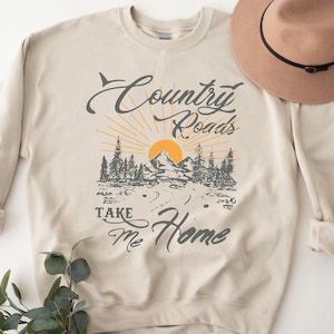 以下が含まれることがあります： 山と太陽のグラフィックプリントが施されたベージュのトレーナー。"Country Roads Take Me Home"の文字がプリントされています。