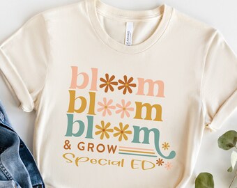 Puede incluir: Una camiseta beige con el texto "bl**m bl**m bl**m & GROW Special ED**" en letras de colores con acentos de flores.