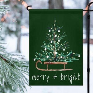Peut inclure: Un drapeau de jardin de Noël vert avec une illustration à l'aquarelle d'un sapin de Noël décoré sur une luge. Les mots "merry + bright" sont écrits en blanc en bas du drapeau.
