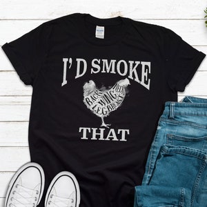 May include: Black t-shirt with a white graphic of a chicken with the words "I'd Smoke That" printed on it. The chicken is labeled with different parts of the bird, such as "back", "wing", and "leg".