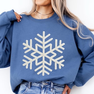 May include: Indigo blue sweatshirt featuring a large, cream-colored snowflake design. The long-sleeved crewneck top is paired with light blue jeans. The text "INDIGO BLUE" is printed in a bold font at the bottom right corner of the image.