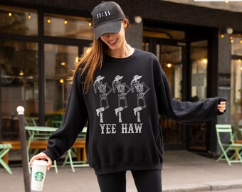 May include: Black sweatshirt with a white graphic of three skeleton cowboys and the text "YEE HAW". The model is wearing a black baseball cap with "11:11" on it, black leggings, and white sneakers. The word "BLACK" is in a white box in the top right corner.