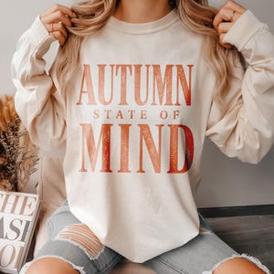 May include: An ivory long-sleeve sweatshirt featuring the text "AUTUMN STATE OF MIND" in a gradient orange and red colour with a speckled texture. The sweatshirt is paired with light blue denim jeans that have large distressed holes at the knees.