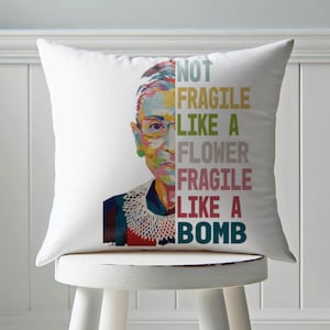 May include: White square pillow featuring a colorful portrait of a person with glasses and a lace collar. The pillow has the text "NOT FRAGILE LIKE A FLOWER FRAGILE LIKE A BOMB" in various colors. The pillow is on a white stool.