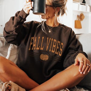 May include: A dark chocolate brown crewneck sweatshirt featuring the text FALL VIBES in a distressed gold font above a graphic of a pumpkin. The long-sleeved garment has a ribbed collar, cuffs, and hem, and is shown in a dark chocolate color.