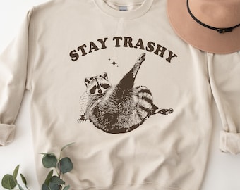 May include: A beige sweatshirt with the text "STAY TRASHY" printed in brown. A cartoon raccoon is printed below the text. The sweatshirt is laying on a white surface with a brown hat and green leaves.