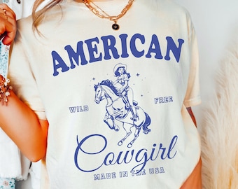 May include: Ivory t-shirt with a blue graphic of a cowgirl riding a horse. The graphic includes the text "AMERICAN WILD FREE Cowgirl MADE IN THE USA".