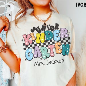 Könnte beinhalten: Elfenbeinfarbenes T-Shirt mit den Worten "Junior Kindergarten" in farbenfrohen Buchstaben im Retro-Stil. Das Shirt hat ein Schachbrettmuster und den Namen "Mrs. Jackson" darunter. Ein lässiges Kleidungsstück für den Alltag.