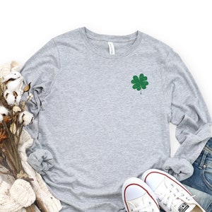 May include: A heather grey long-sleeved t-shirt with a green four-leaf clover embroidered on the left chest.