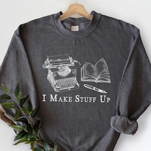 以下が含まれることがあります： タイプライター、開いた本、ペンの白いグラフィックが特徴の濃いヘザーグレーのスウェットシャツ。「I MAKE STUFF UP」の文字が下に印刷されています。「DARK HEATHER」の文字が右下に印刷されています。