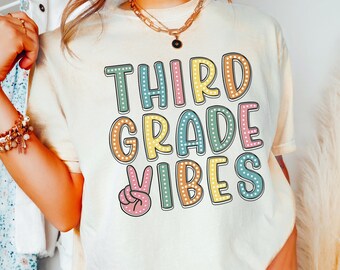 May include: A white t-shirt with the text "THIRD GRADE VIBES" in colorful block letters with a peace sign. The text is outlined in black and has white polka dots.